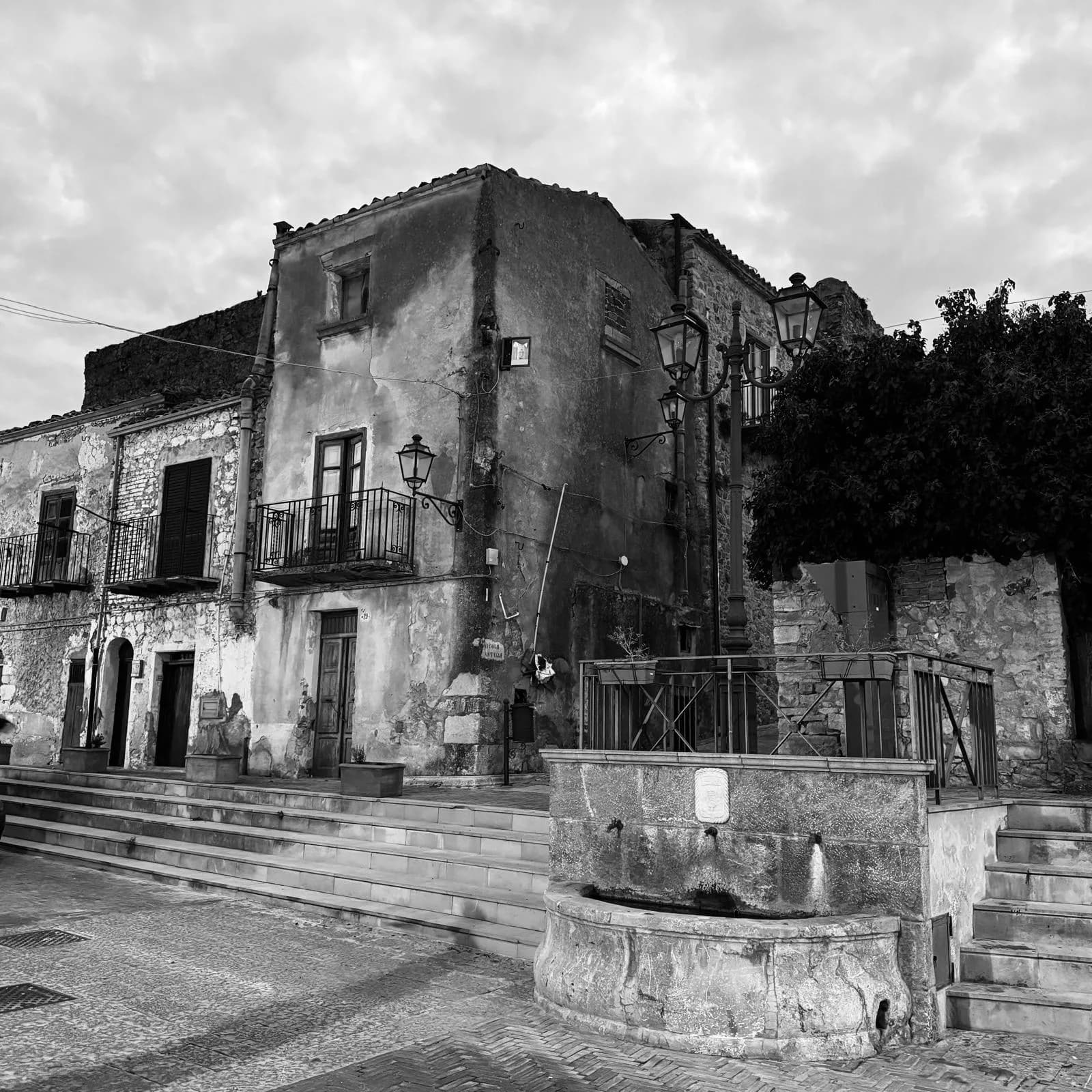 A weathered stone piazza with a fountain, wrought-iron street lamps, and balconied houses in a Madonie village