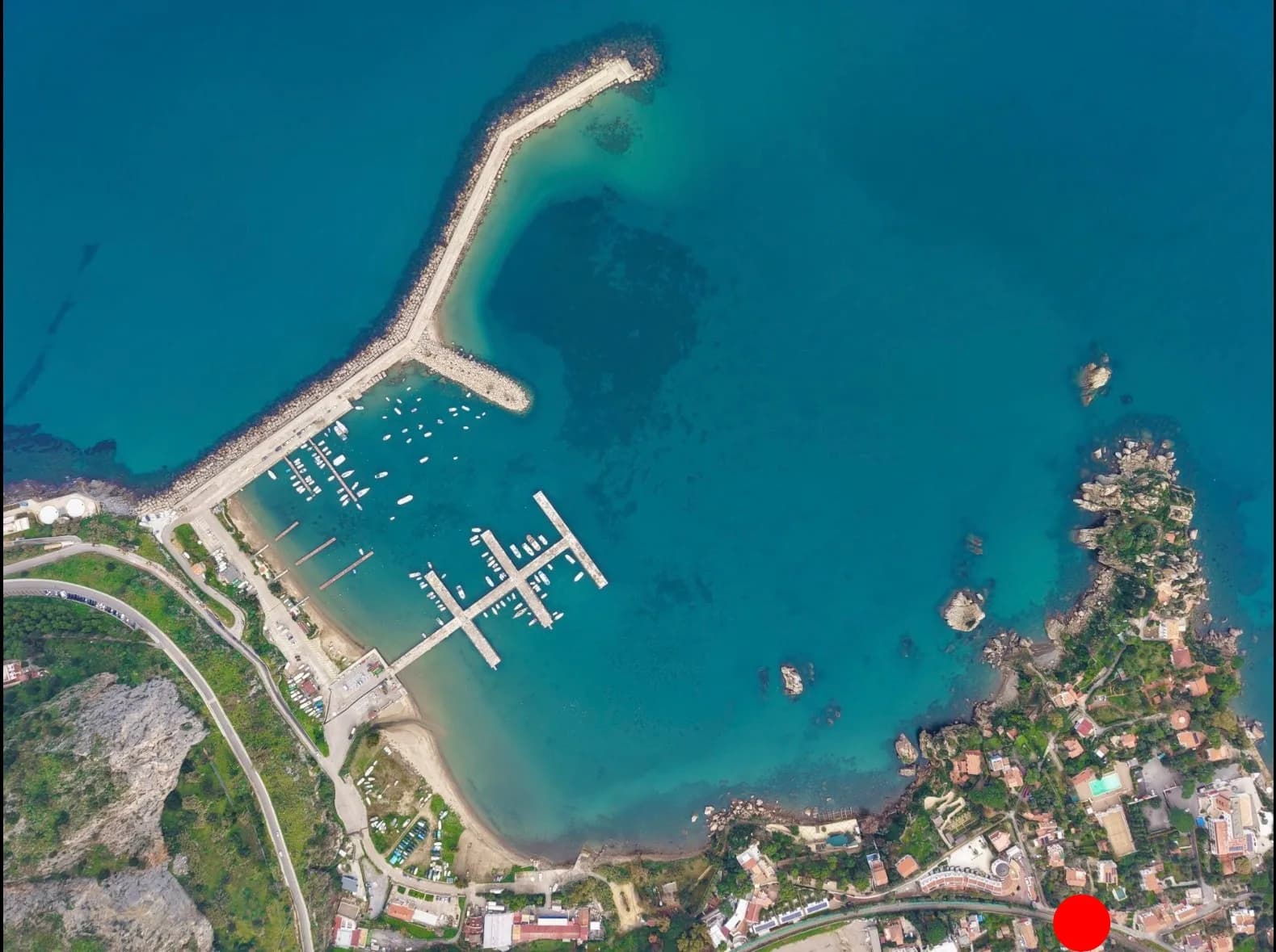 Overhead aerial of Cefalù Marina — the breakwater, moored boats and jetties beside the coastal road, with the property location marked
