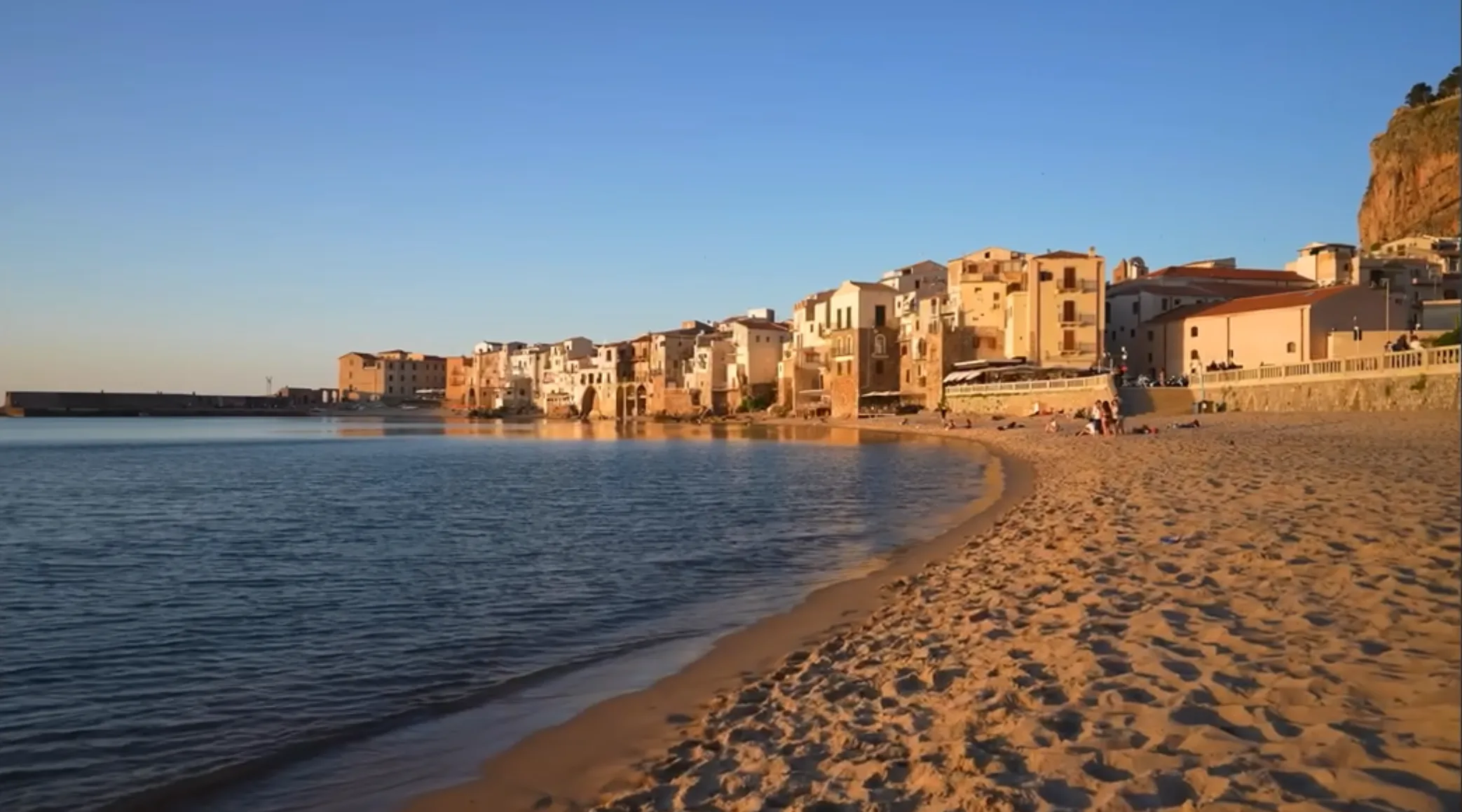 Golden-hour light over the old town and sandy beach of Cefalù, with La Rocca beyond