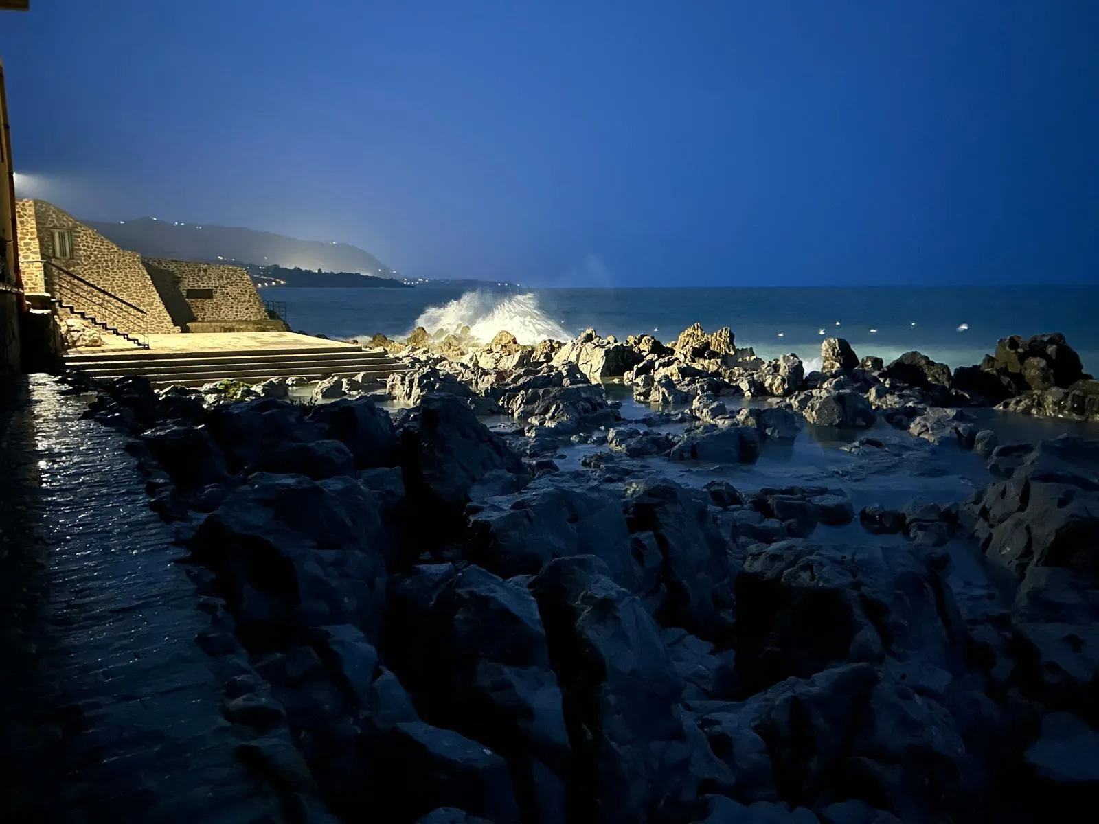 Blue-hour view of the rocky Cefalù coast with waves crashing and mountains in the distance